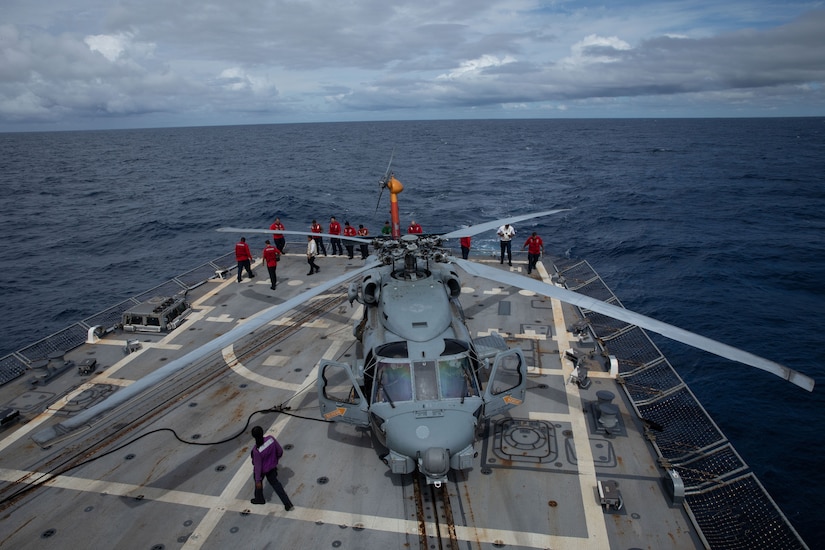 Sailors wearing red, white, green and purple long-sleeve T-shirts, walk around a helicopter on the deck of a ship at sea.