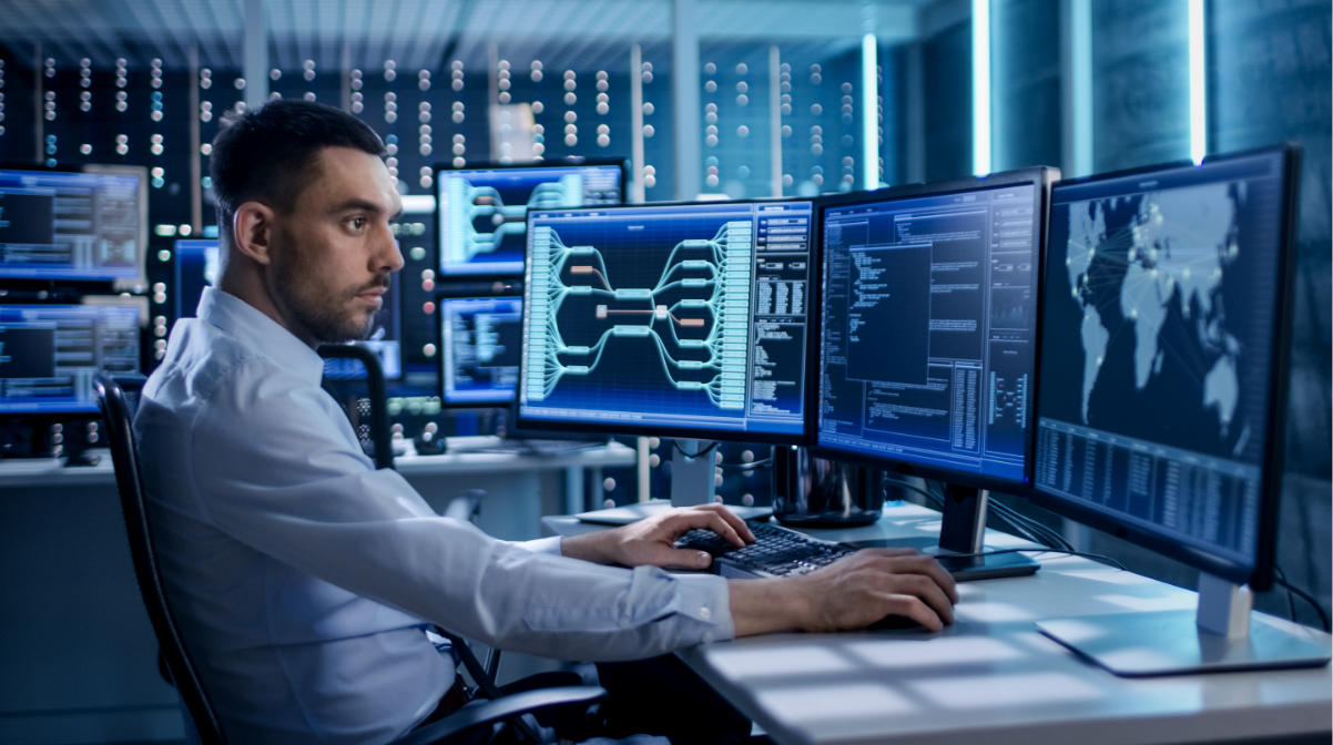 A cybersecurity professional working at his desk in front of three computer screens and with computer screens in the background.
