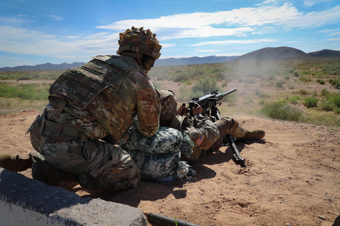 A soldier in tactical gear crouches behind another soldier firing a machine gun in a dirt patch in a field on a sunny day.