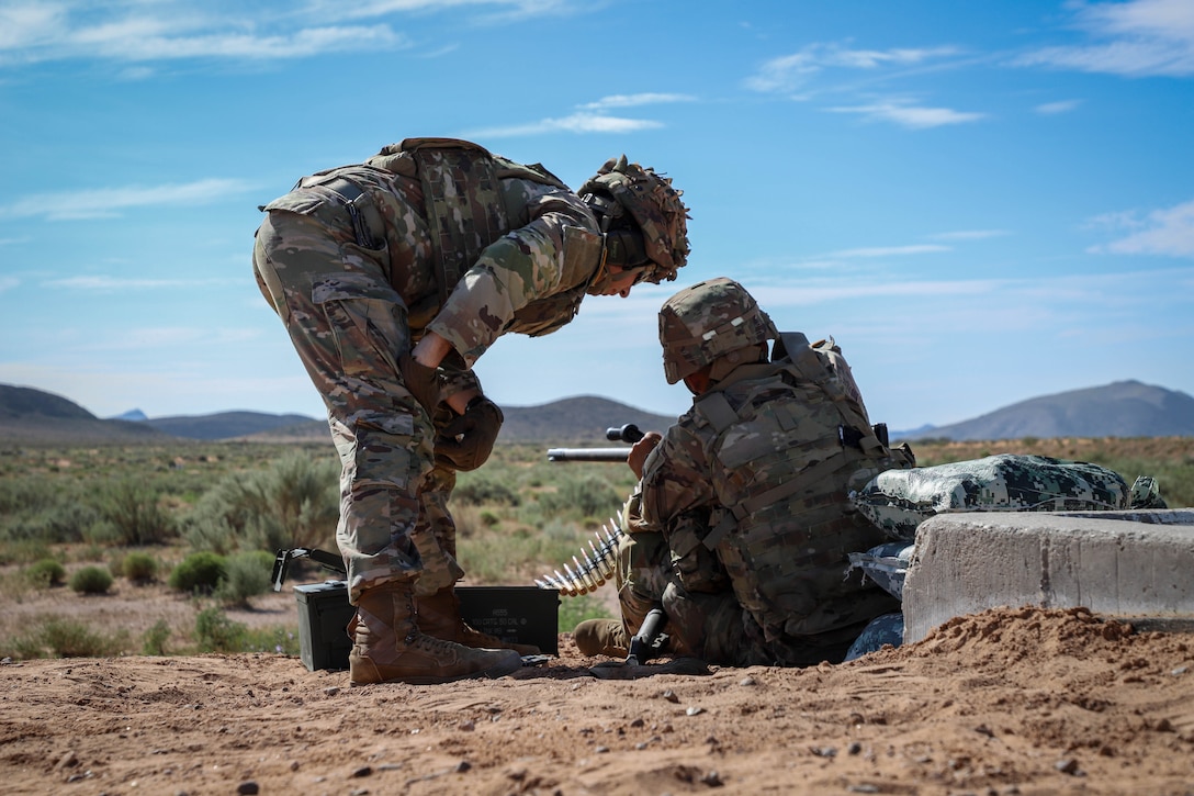 A soldier adjusts a machine gun on the ground as another soldier leans over to observe in a field under blue skies as seen from behind.