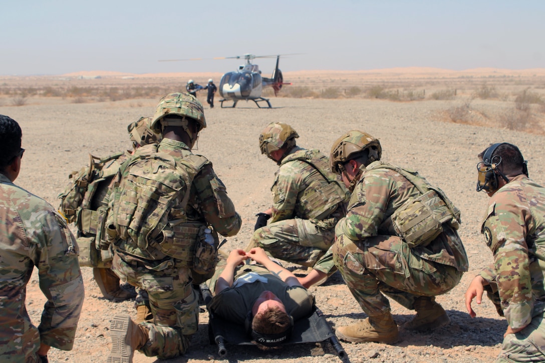 A group of soldiers crouch around a person on a stretcher on sandy terrain with a grounded helicopter in the background under blue skies.