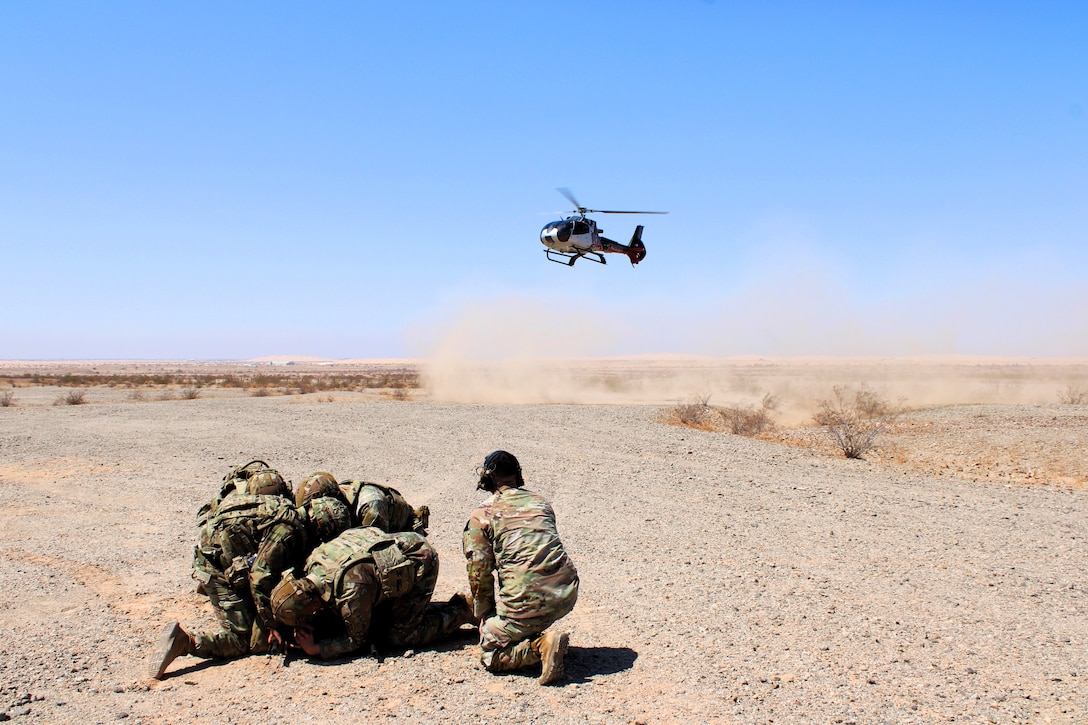A group of soldiers crouch together on  rough, sandy terrain as a helicopter kicks up dust in the distance against a blue, cloudless sky.