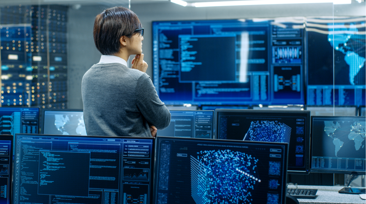A man in a data room with computer screens, standing and looking at data on the large screens.