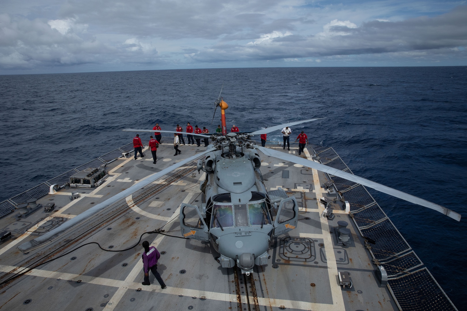 Sailors wearing red, white, green and purple long-sleeve T-shirts, walk around a helicopter on the deck of a ship at sea.