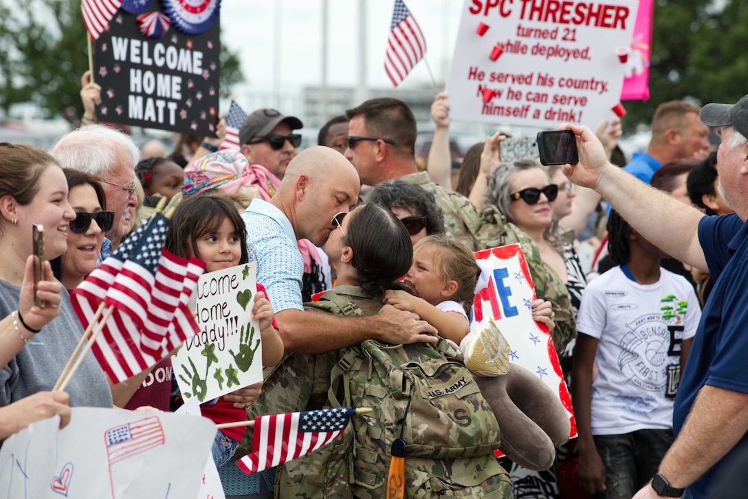 A guardsman kisses a loved one and holds a child in a crowd of people holding colorful signs and flags outside during the day.