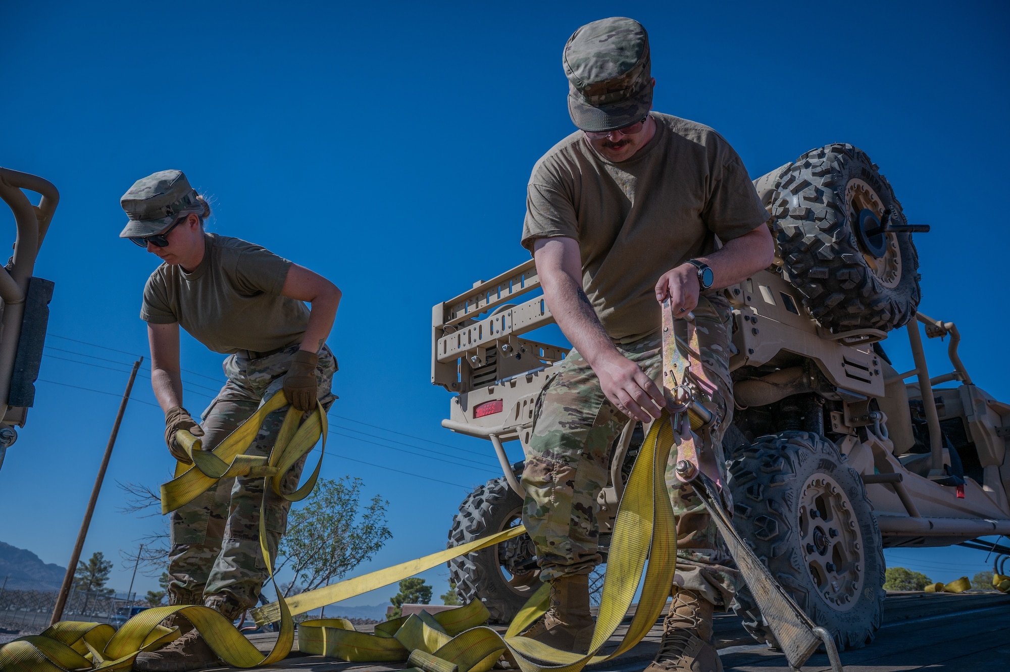 Two Airmen loosed yellow cargo straps aboard a trailer with a utility vehicle in the background.