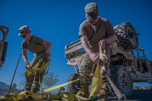 Two Airmen loosed yellow cargo straps aboard a trailer with a utility vehicle in the background.