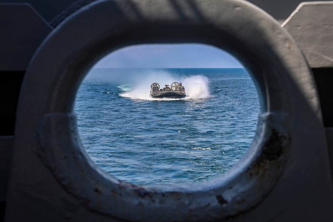 A small boat sails on the ocean during the day toward a ship as seen through an opening on the ship.