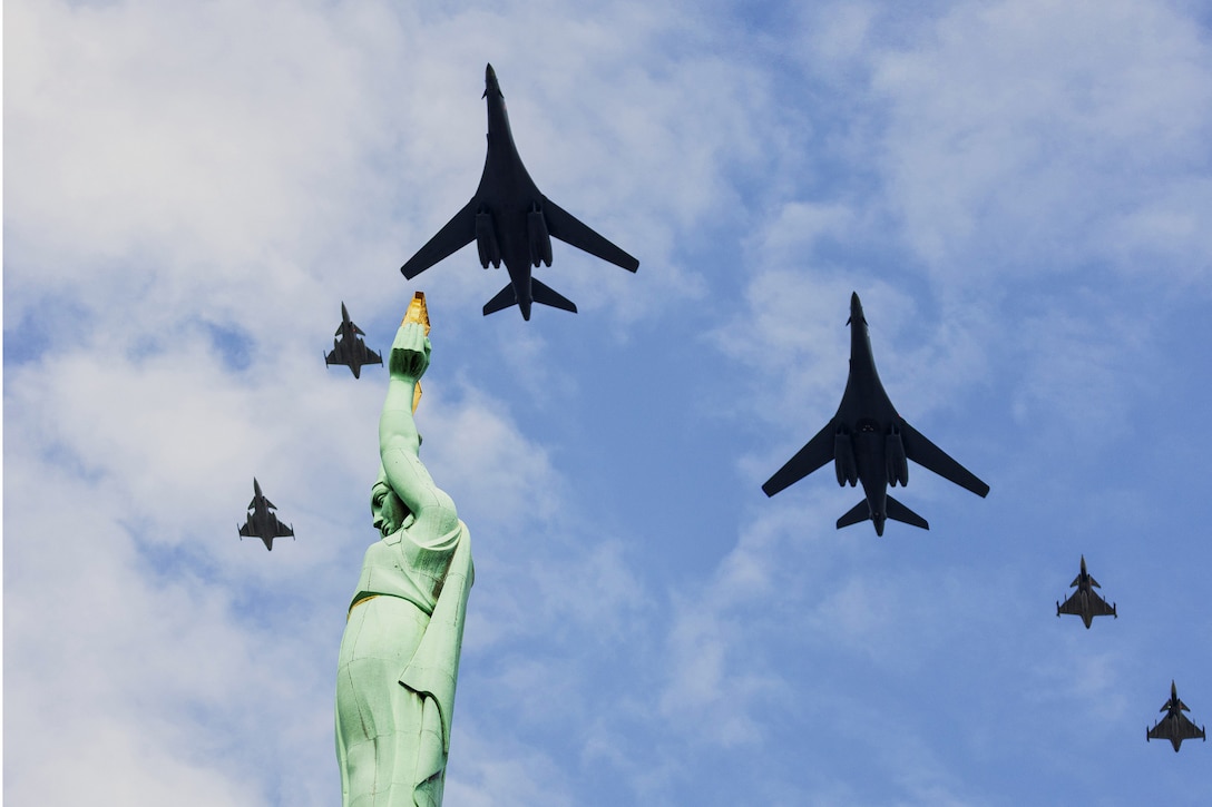 Six aircraft fly over a green and gold statue in a blue sky with clouds as seen from the ground.