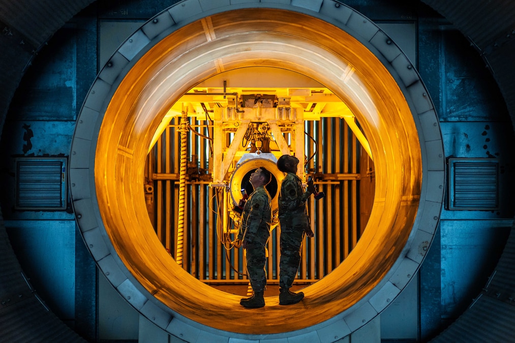 Two airmen stand back-to-back in a large metal tube structure bathed in yellow light while facing opposite sides.