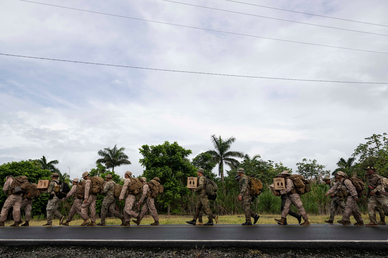 A group of service members walk down a road next to a jungle carrying supplies.