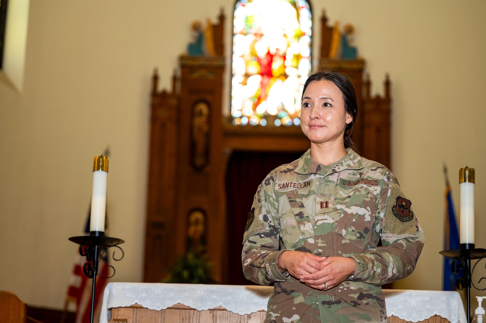 Woman in uniform holds hands together and is standing in front of the alter inside the chapel. She is looking to the side. She is by herself.