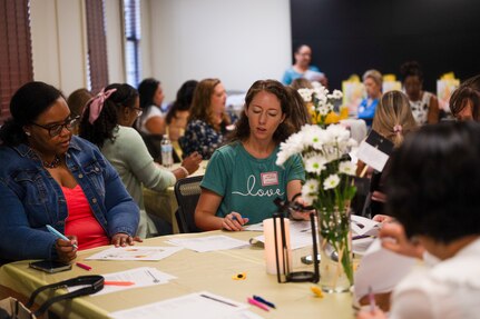 Women sitting at a table inside a building. On the tables there are flower arrangements and papers. One lady is holding a pen and writing and the other lady is looking down reading the paper.
