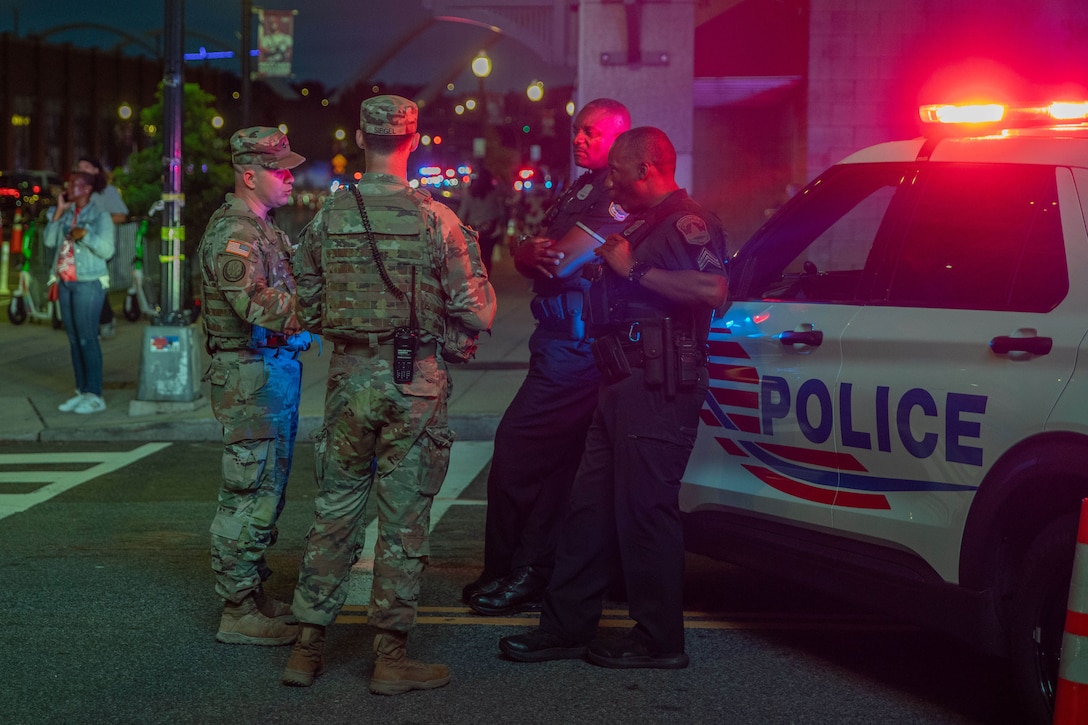 Two soldiers stand in the middle of a street at night and speak with two police officers standing in front of a police vehicle.
