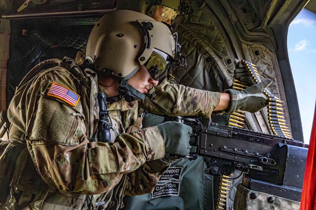 Two soldiers in helmets work together to operate a machine gun positioned out of an aircraft.