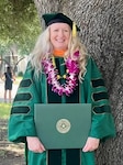 Female Soldier standing in front of a tree dressed in college graduation regalia and holding her diploma.