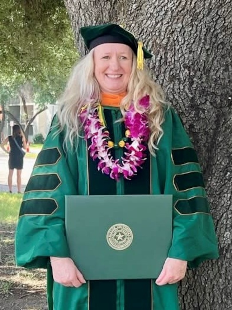 Female Soldier standing in front of a tree dressed in college graduation regalia and holding her diploma.