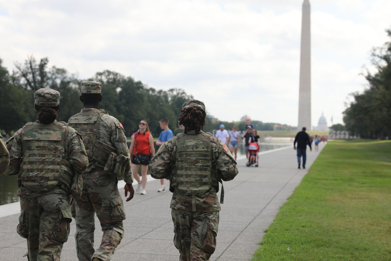 Three soldiers walk together down a pathway. The Washington Monument and the U.S. Capitol are in the background.
