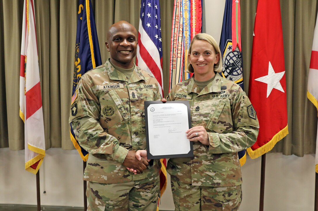 Two soldiers pose in front of standing flags while holding a certificate and shaking hands.