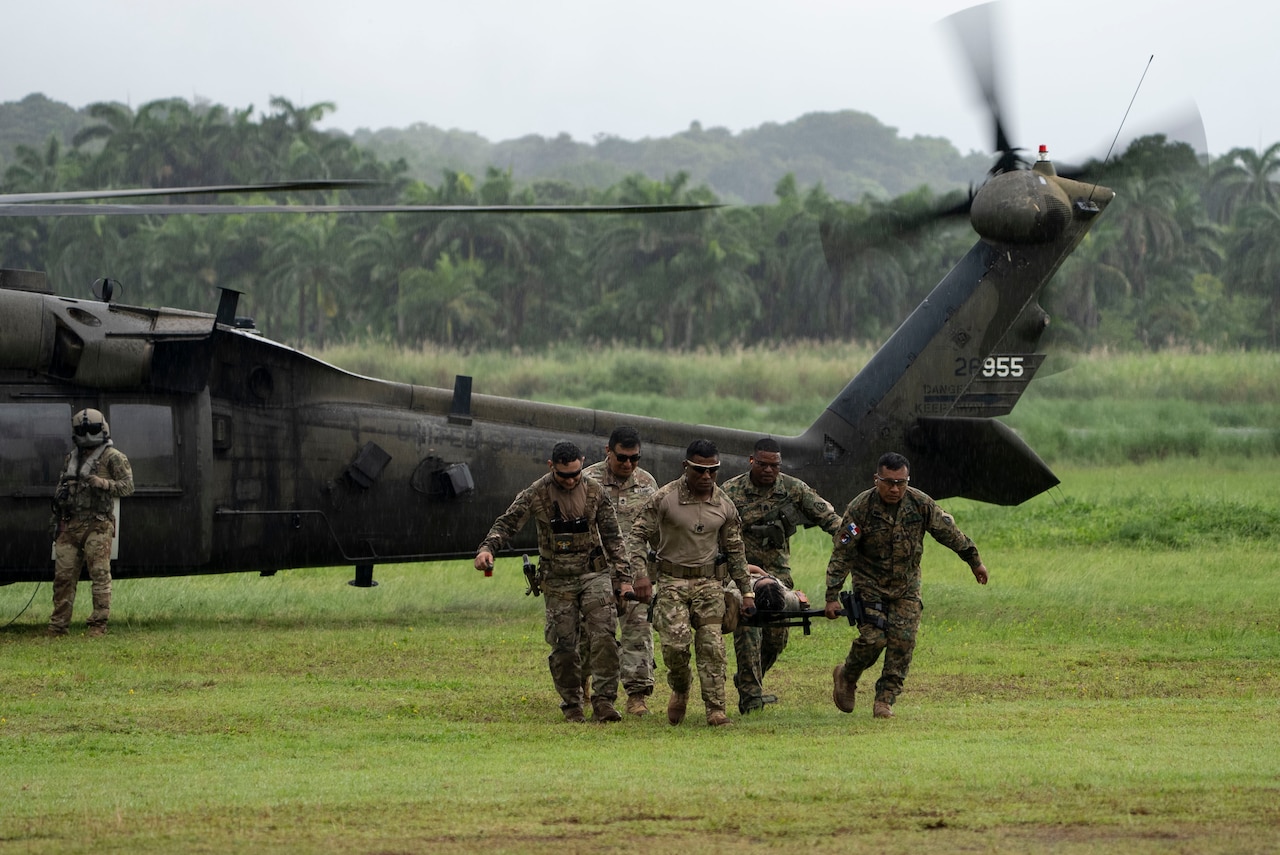 A group of service members carries a stretcher away from a parked helicopter in a jungle clearing as a pilot watches on.