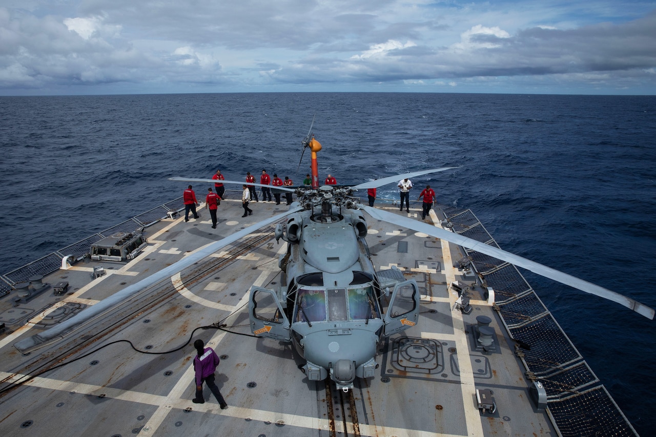 Sailors wearing red, white, green and purple long-sleeve T-shirts, walk around a helicopter on the deck of a ship at sea.
