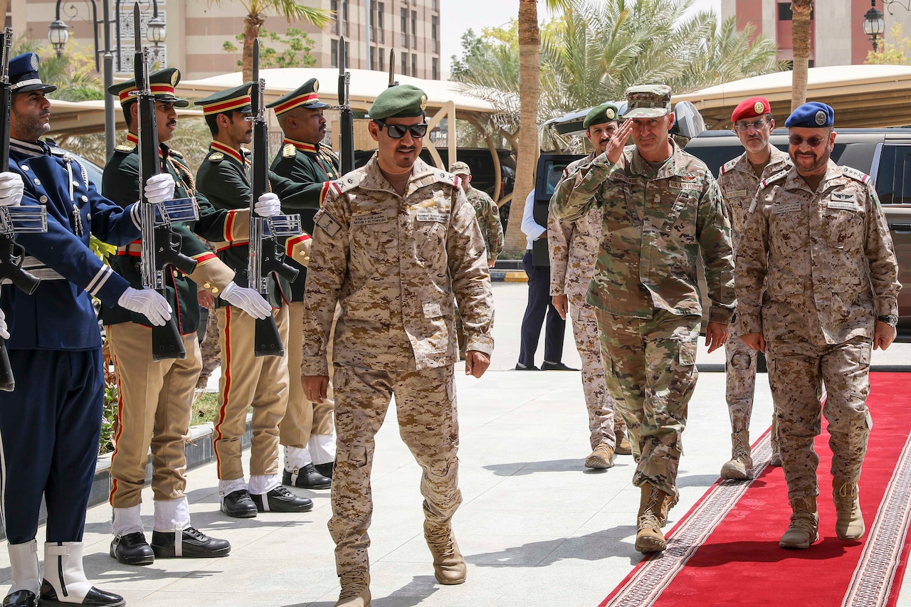 An airman salutes while walking with Saudi Arabian armed forces service members on a red carpet outdoors, as other Saudi Arabian troops stand at attention holding rifles upright.
