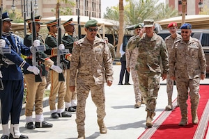 An airman salutes while walking with Saudi Arabian armed forces service members on a red carpet outdoors, as other Saudi Arabian troops stand at attention holding rifles upright.