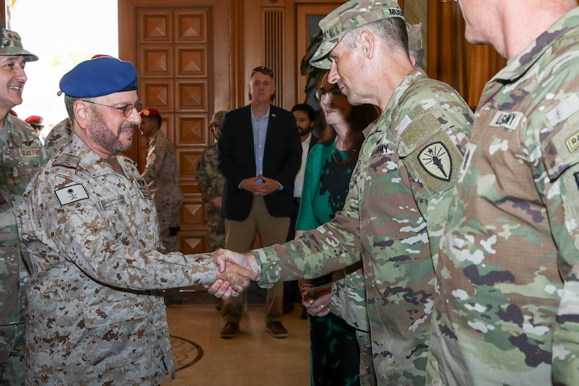 Two people wearing camouflage military uniforms shake hands indoors as other people, some in military uniforms and some in civilian clothes, stand and watch.