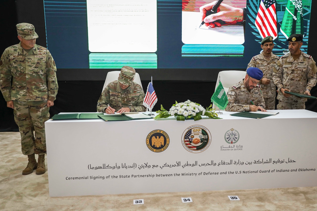 Two people in camouflage military uniforms sign papers while seated at a table indoors as three others in military uniforms stand and look on.