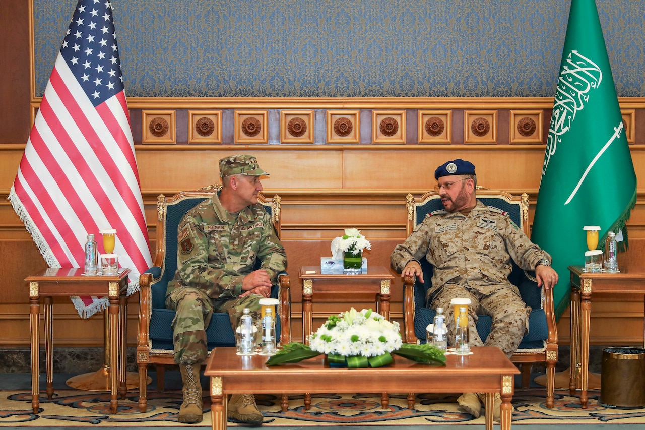 Two people wearing camouflage military uniforms sit in chairs next to each other indoors and speak, with an American flag and a Saudi Arabian flag also visible.