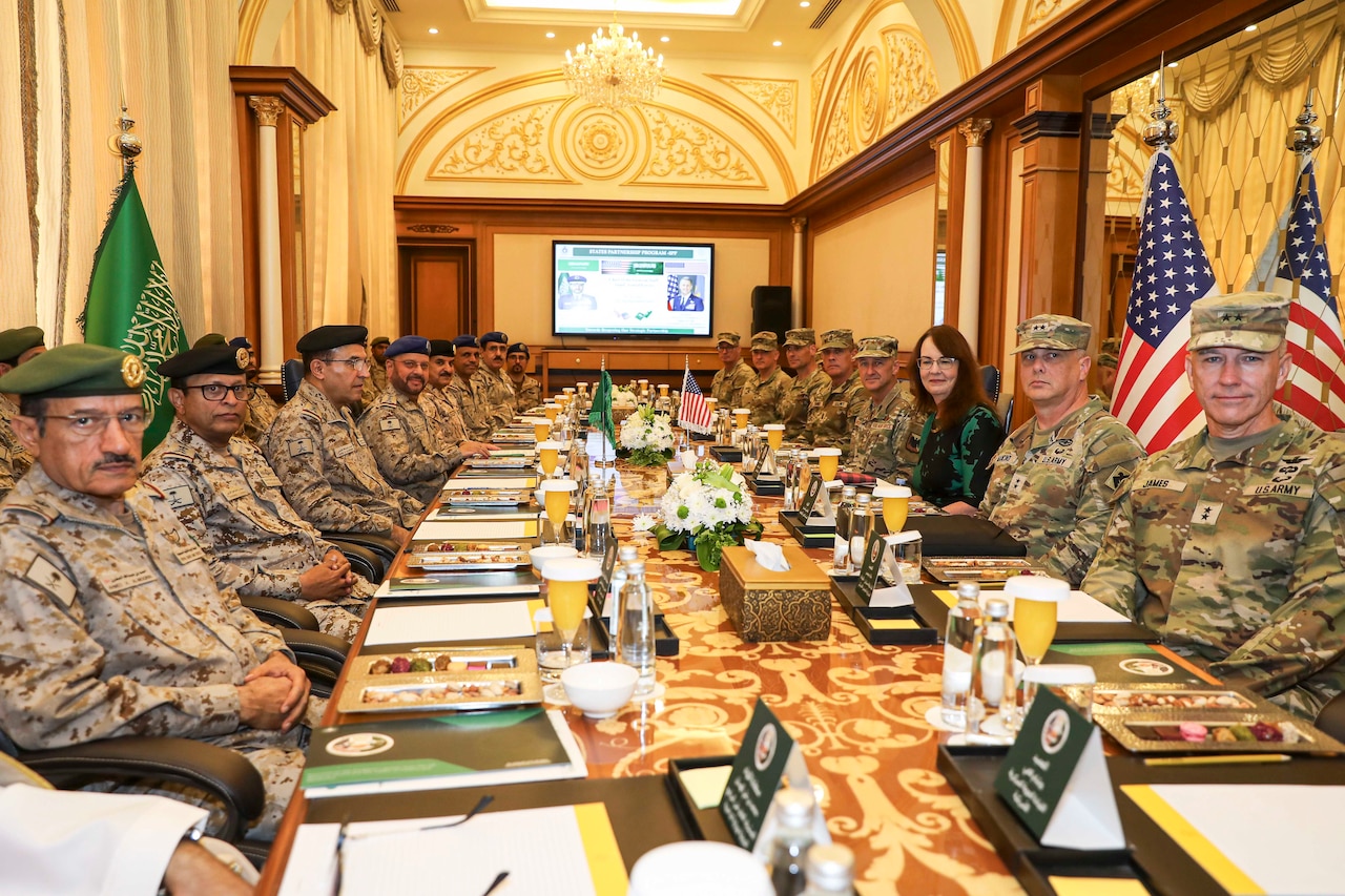 People in camouflage military uniforms and a person in civilian clothes sit on each side of a long table, with a Saudi Arabian flag and two American flags on poles near the walls of an ornate room, with a display screen mounted on another wall.