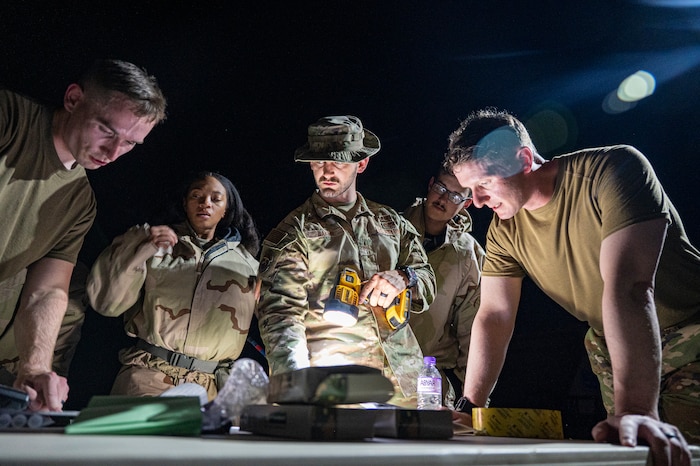 386th ECES Airmen huddle around a table as some members are changing out of MOPP gear and others moving into gear as they hear a briefing