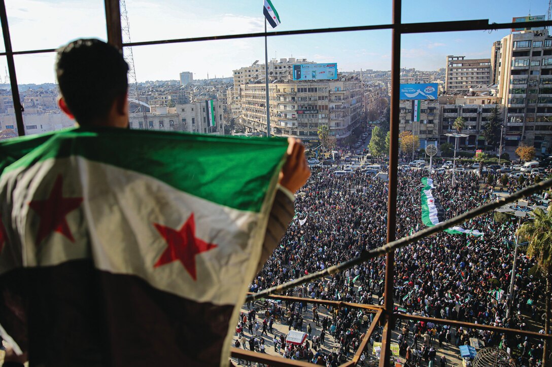 A person draped in the Syrian opposition flag looks out over a large protest in a city square, where a massive crowd gathers, many carrying similar flags. The scene is framed by tall buildings and city streets filled with people.