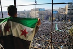A person draped in the Syrian opposition flag looks out over a large protest in a city square, where a massive crowd gathers, many carrying similar flags. The scene is framed by tall buildings and city streets filled with people.