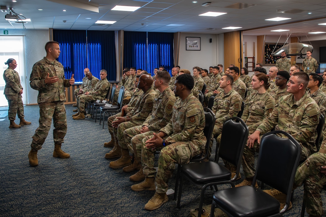An Air Force colonel stands in front of and speaks to a room full of Airmen