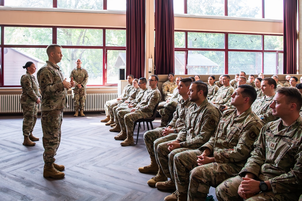 An Air Force colonel stands in front of and speaks to a room full of Airmen