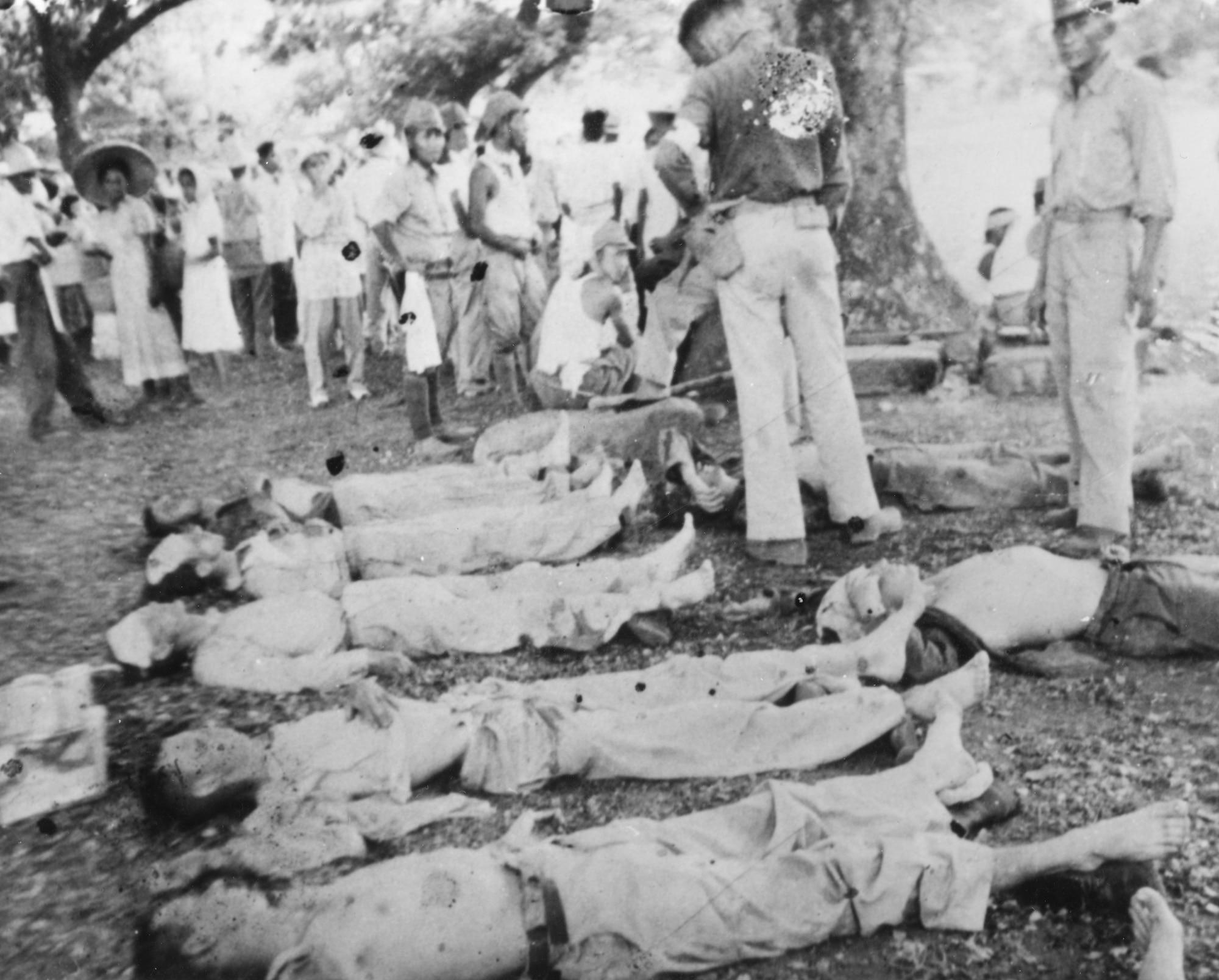 A row of murdered POWs lay on the ground with Japanese soldiers and prisoners standing around them.