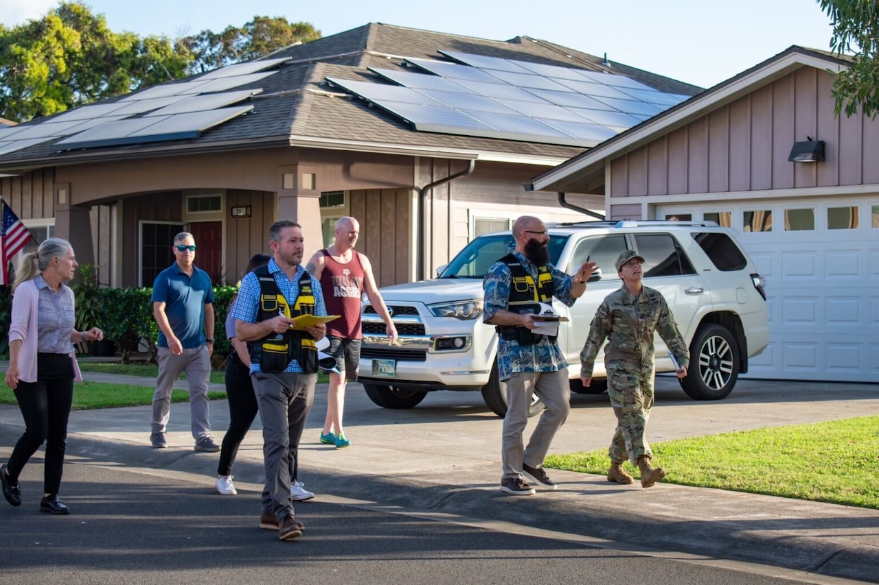 A woman wearing a camouflage military uniform walks down a neighborhood sidewalk with a bearded man dressed in casual attire and a black vest walking alongside her. There is also another man in similar attire, as well as two other men and a woman also dressed casually, walking behind them. There is a vehicle in a driveway and a house behind them.