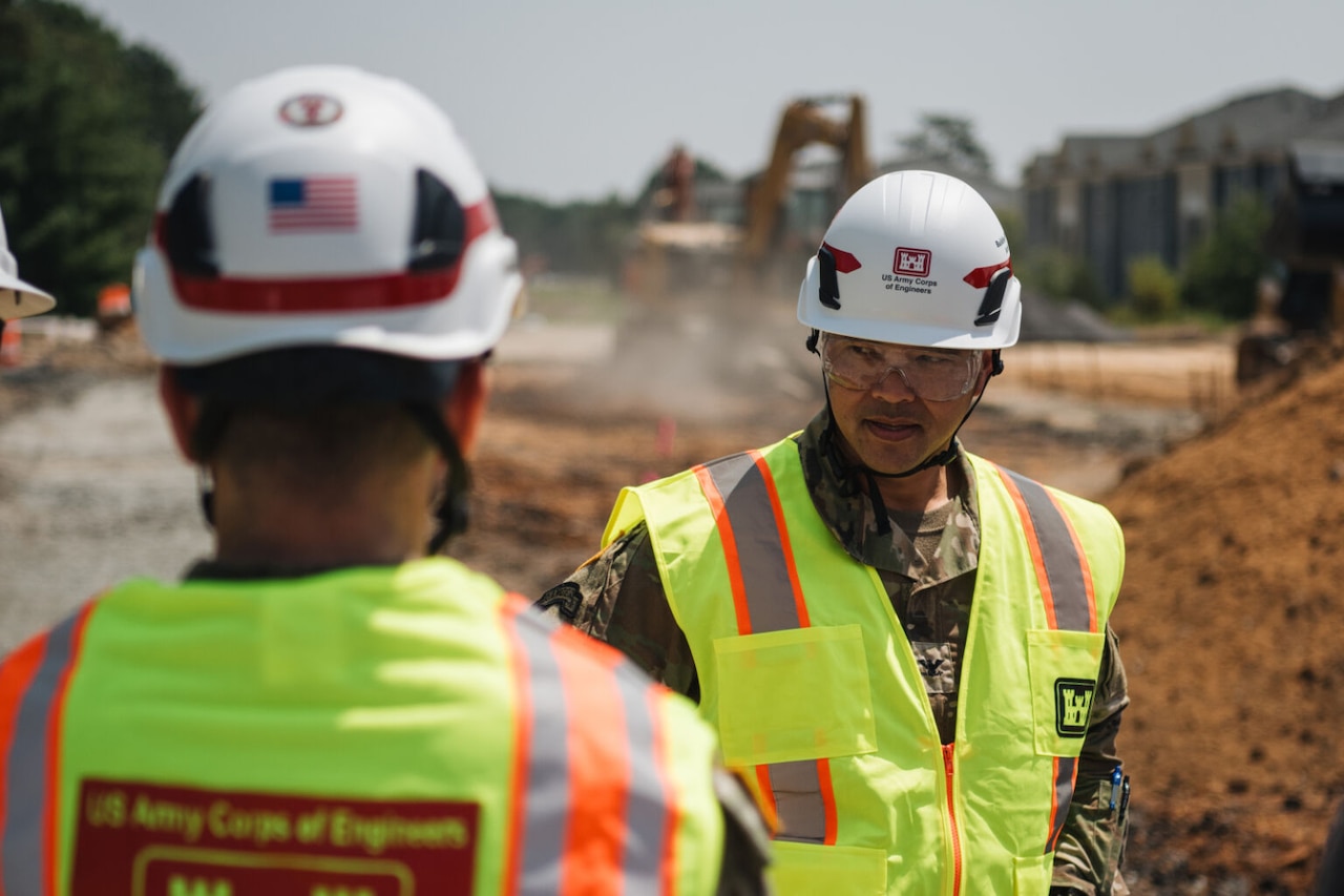 A man in a camouflage military uniform wearing a reflective safety vest and a construction helmet looks to the left. A man in similar attire stands in front of him, and an excavator behind him moves dirt on a construction site.