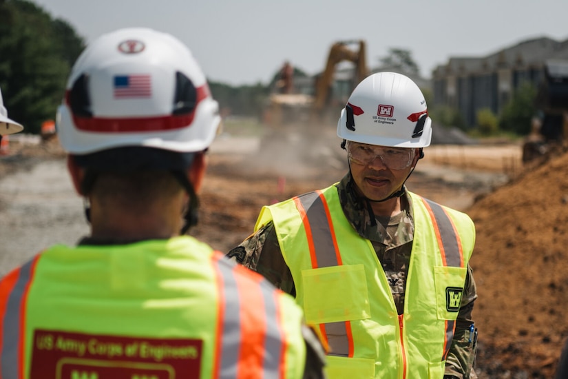A man in a camouflage military uniform wearing a reflective safety vest and a construction helmet looks to the left. A man in similar attire stands in front of him, and an excavator behind him moves dirt on a construction site.