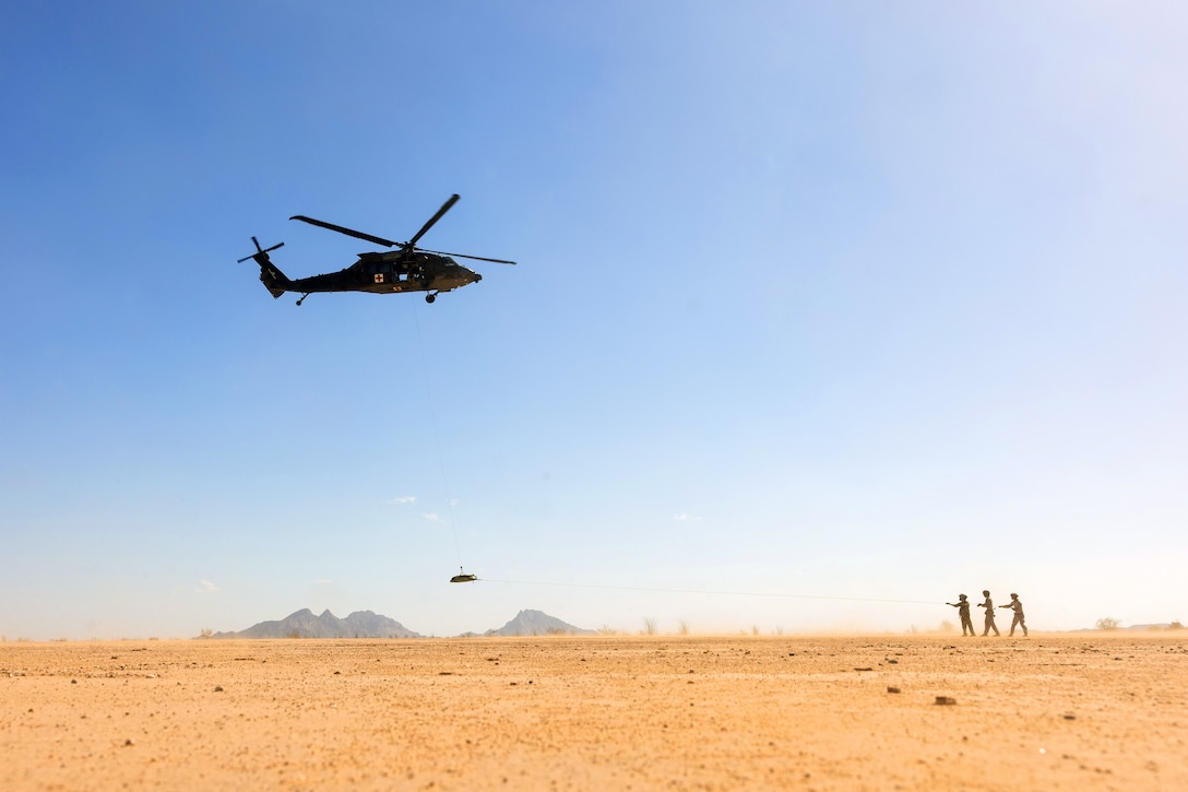 Three service members stand in a desert like setting as a helicopter hovers and a simulated casualty hangs from it.