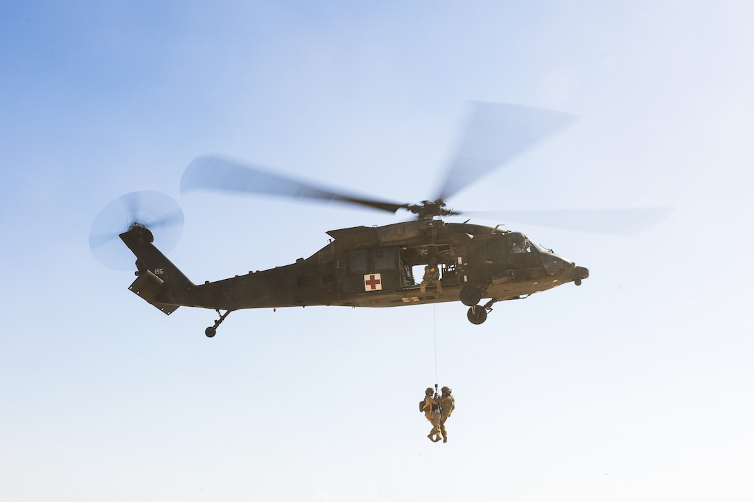 A soldier wearing a harness and a training mannequin hang from a helicopter in flight against a blue sky.
