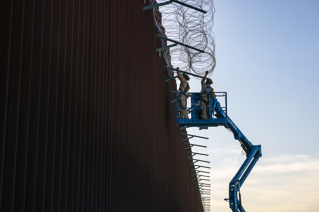 Two Marines standing on a raised blue platform hold up concertina wire against a tall wall.