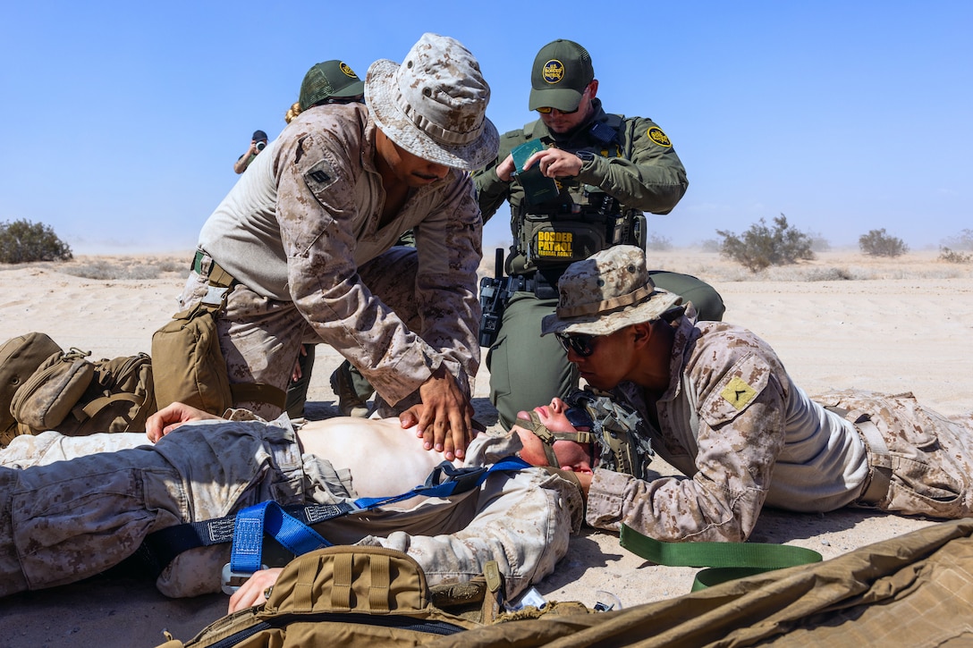 Four Marines kneel around another Marine and simulate giving CPR under blue skies.