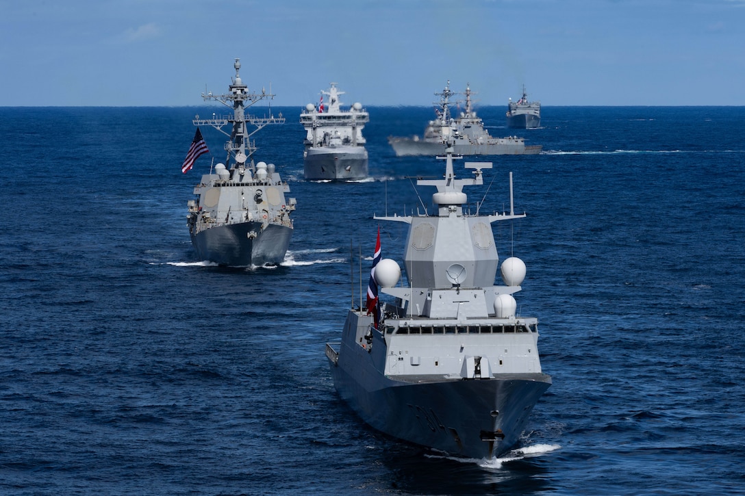 Five ships with flags sail in formation on the ocean during the day.