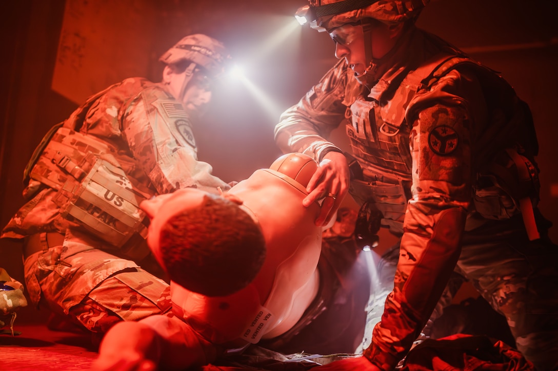 Two soldiers in tactical gear wearing helmets with flashlights kneel and turn a training mannequin to the left in a dark room bathed in red light.
