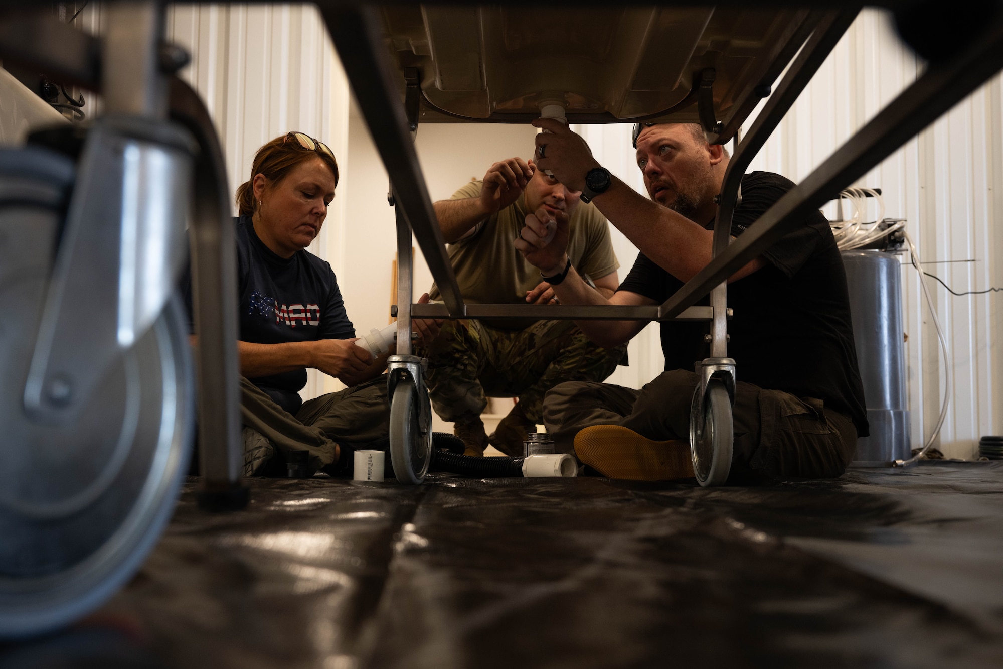 Danielle Wilk, Air Force Mortuary Affairs Operations port mortuary branch chief, Senior Master Sgt. Alfredo Jauregui, AFMAO operations support division superintendent, and Matthew Metschke, AFMAO case management branch chief, build a drainage system under an embalming table at Grayling Army Airfield, Michigan, Aug. 9, 2025. The system was a key part of the Expeditionary Mortuary AFMAO members created during Northern Strike 25-2. (U.S. Air Force photo by Senior Airman Madeline Baisey)