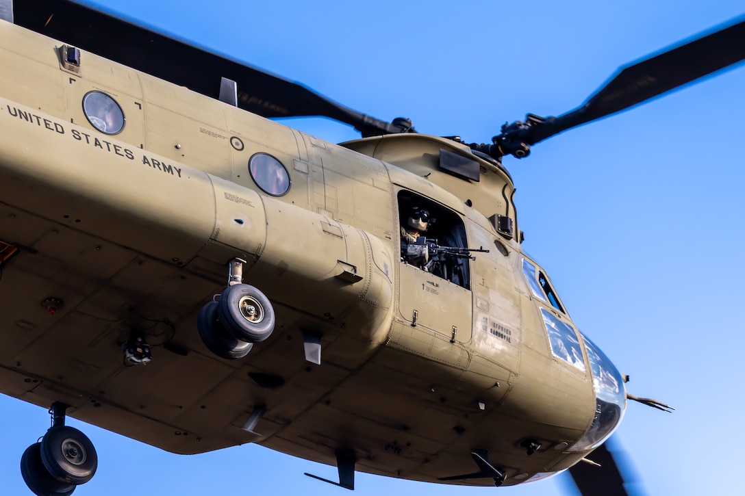 A soldier holding a service weapon leans out of a helicopter in flight under blue skies.