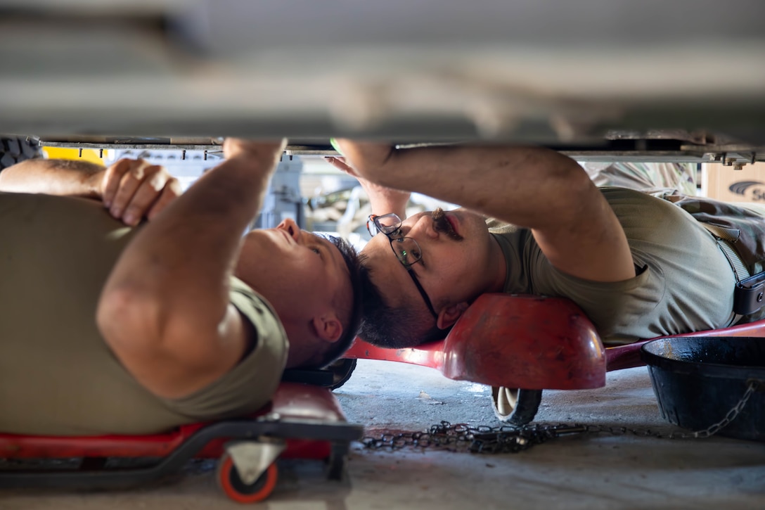 Two guardsmen lay on their backs on red garage creepers and reach up into a vehicle.