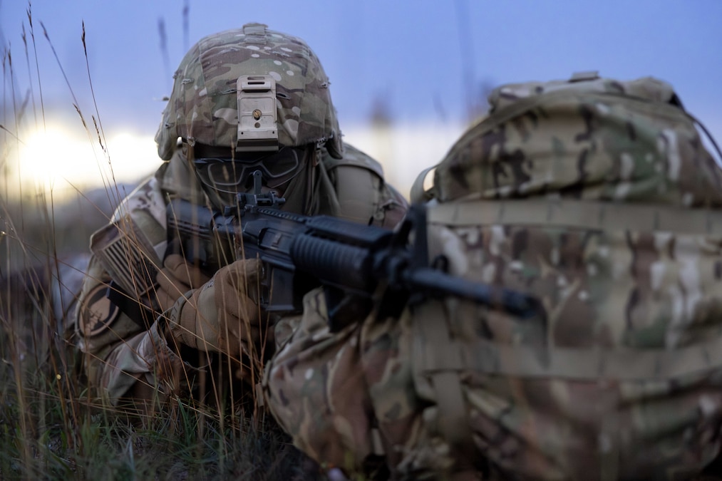 A soldier in tactical gear leans a service weapon on a backpack to aim to the right while laying in a field during the day.